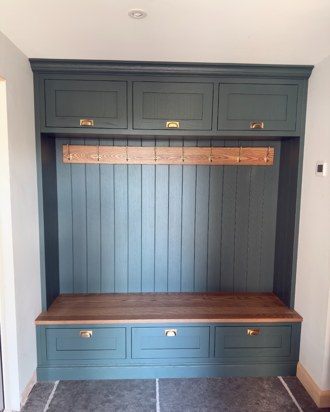 Dark green boot room with oak bench seat, brass cup handles, tongue-and-groove panelling, and coat hooks